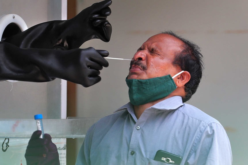An Indian man in a blue shirt and face mask around his neck grimaces as a swab goes up his nose.
