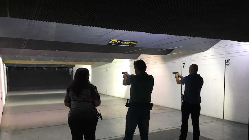 Three people face targets at a shooting range, two of them with their guns out and ready to fire