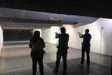 Three people face targets at a shooting range, two of them with their guns out and ready to fire