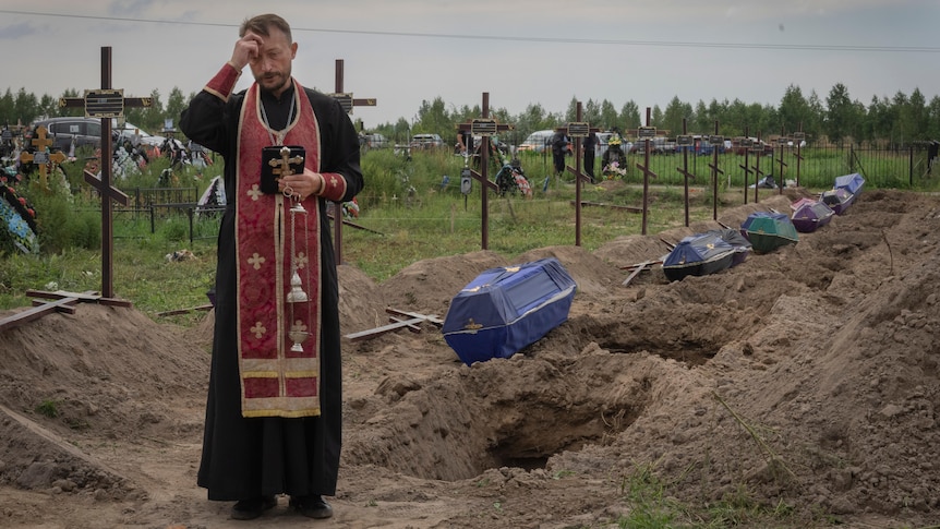 An orthodox priest in red and black gear prays in front of a row of open graves with covered coffins waiting next to them 
