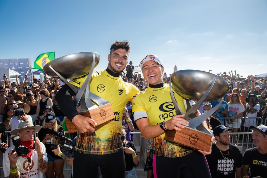 A man and a woman in yellow wetsuits smile at the camera while each holding trophies