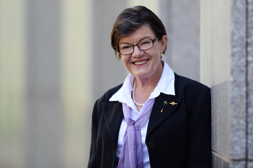 Cathy McGowan smiles at the camera while standing against a wall.