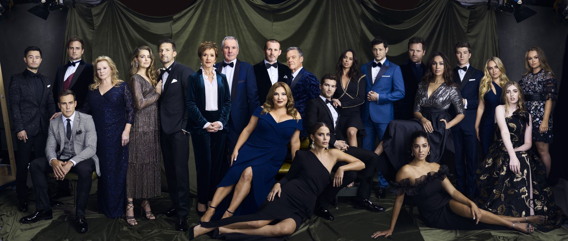 The main cast members of Neighbours, wearing suits and formal dresses and posing for a photo in a studio.