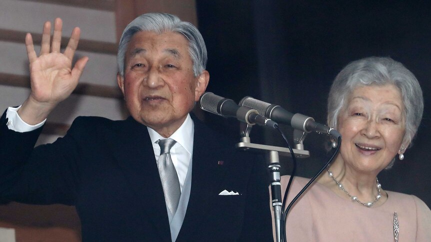 Emperor Akihito standing in front of a microphone and waving, with his wife Empress Michiki standing and smiling next to him