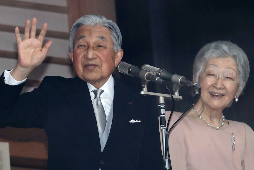 Emperor Akihito standing in front of a microphone and waving, with his wife Empress Michiki standing and smiling next to him