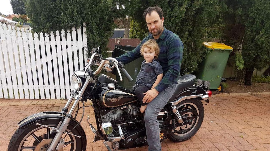 A man sitting on a stationary motorbike with a toddler perched in front of him.