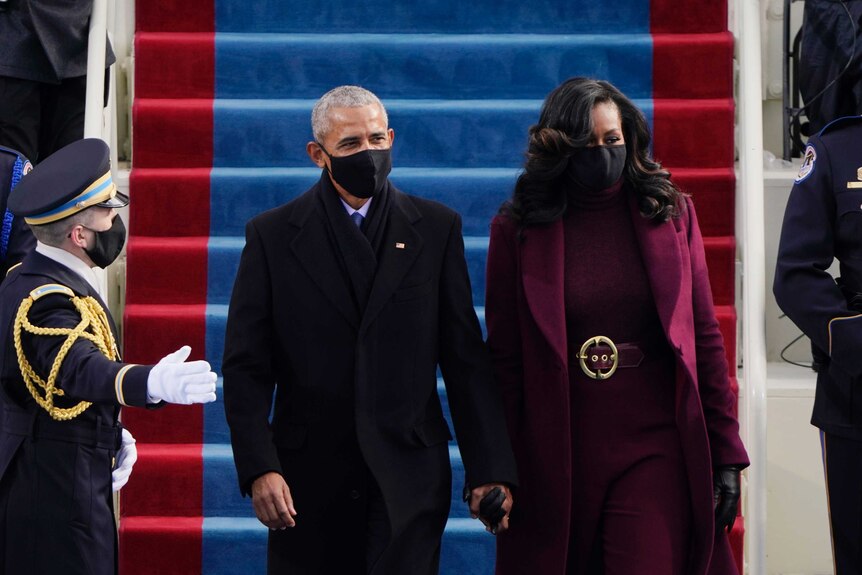 A man wearing a black mask and suit holds hands with a woman wearing a mask and burgundy suit.