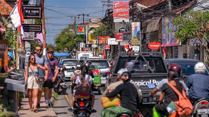 Tourists walk on a narrow footpath as scooters and cars drive on a congested road beside them.