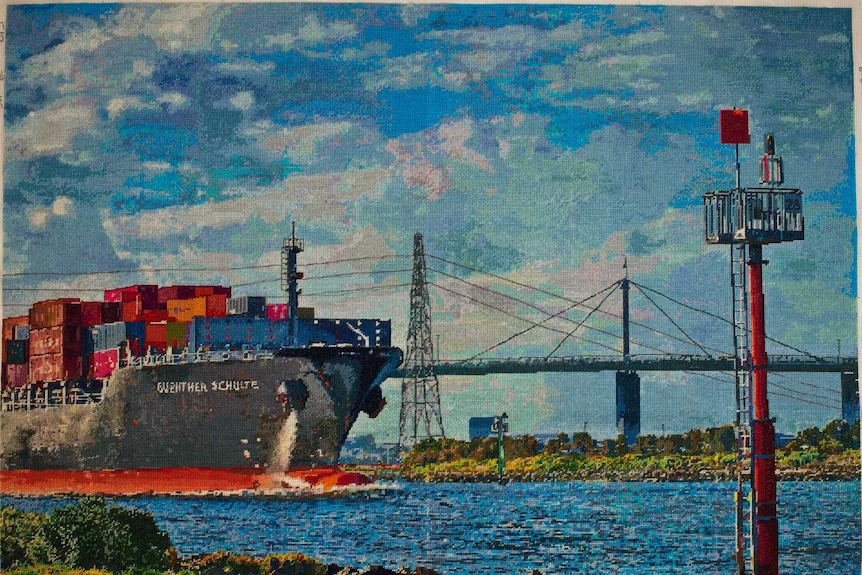 Needlepoint work showing a large shipping container freight boat sailing down a river, with a cloudy sky above it