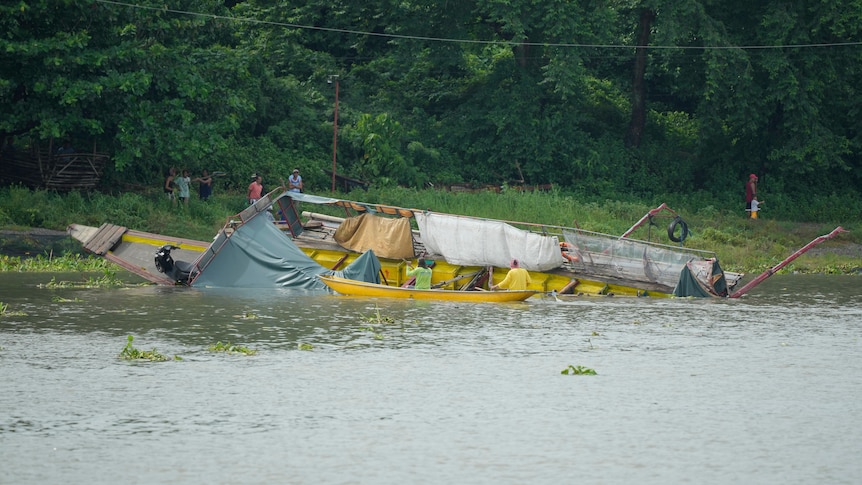 A partially overturned boat washed up on the bank of a body of water.
