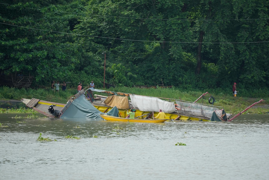 A partially overturned boat washed up on the bank of a body of water.
