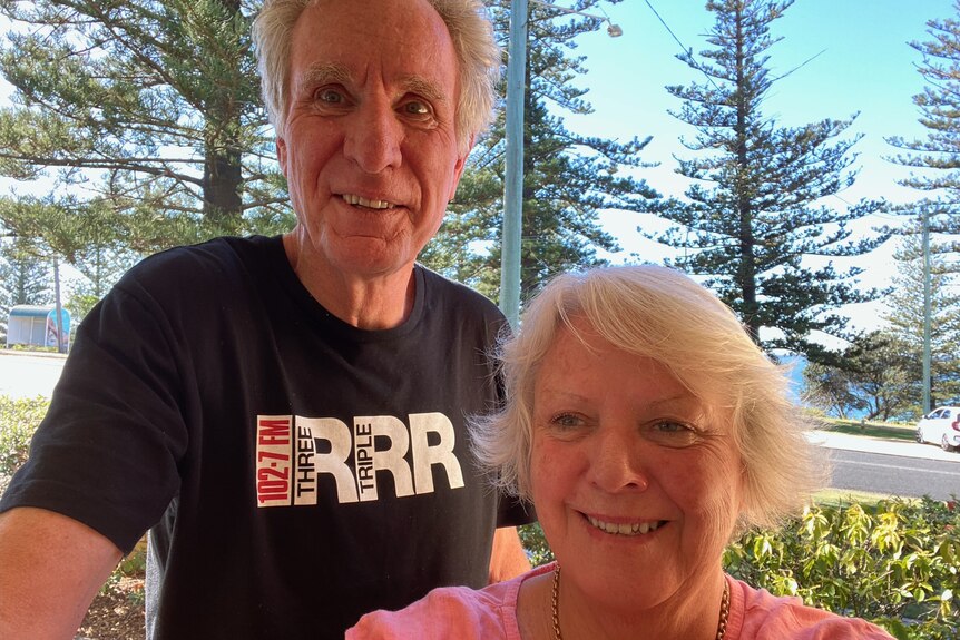 A woman in her sixties standing next to her husband with pine trees and the ocean in the background.