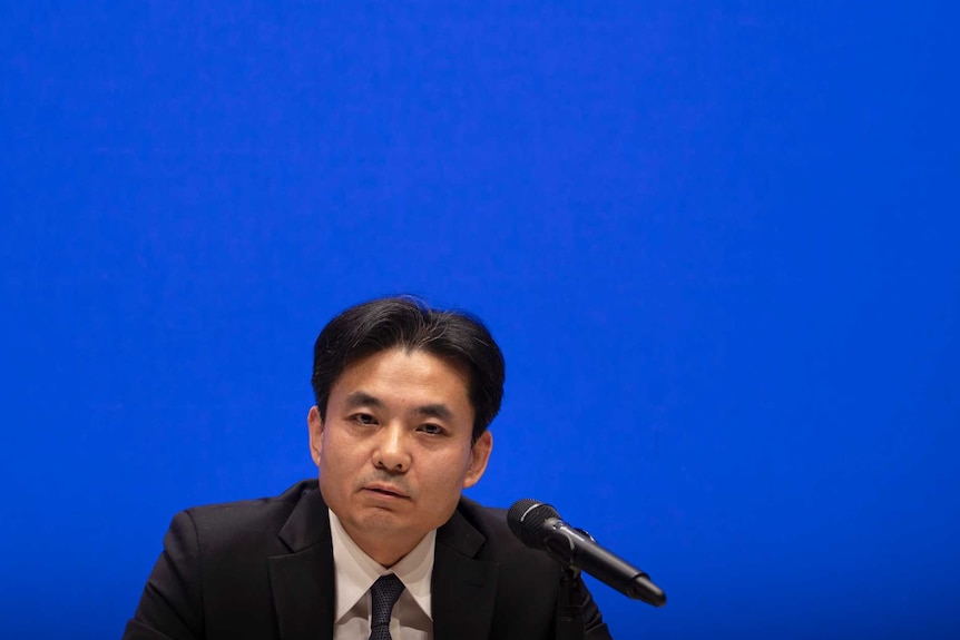 Yang Guang wears a suit and speaks in front of a large blue sign
