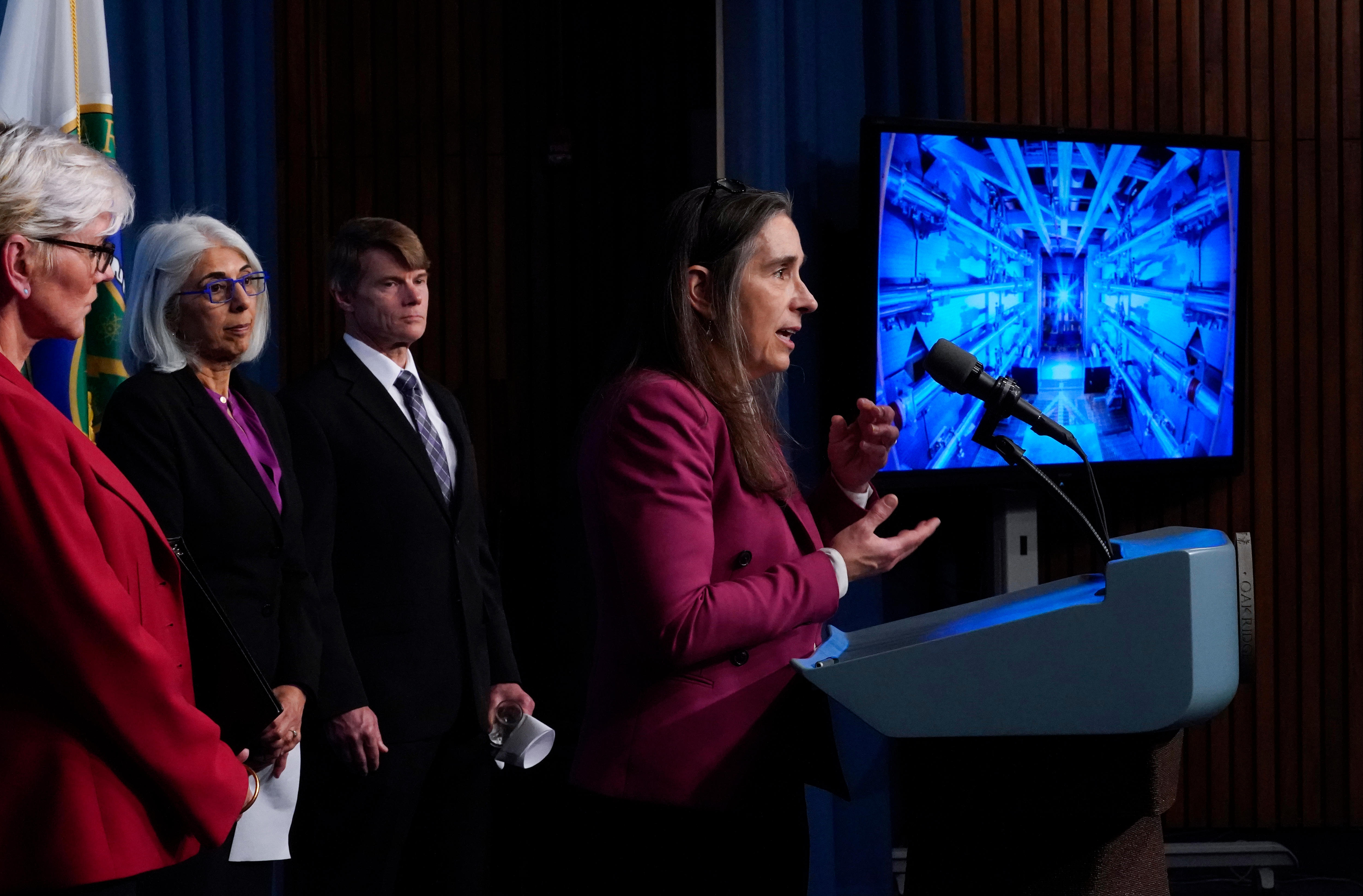 A woman in magenta blazer speaks at podium in front of screen with blue image