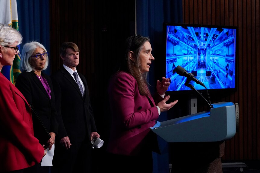 A woman in magenta blazer speaks at podium in front of screen with blue image