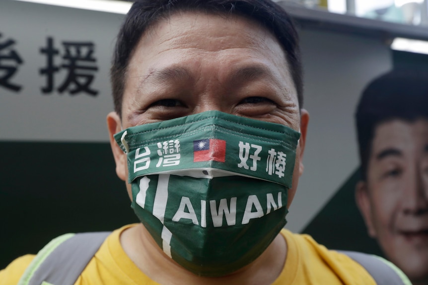 a man wears a green mask with words including Taiwan and the Taiwanese flag on it as he looks at the camera