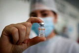 You view a woman holding a tiny vial of the Sinovac vaccine between her thumb and index finger in the centre of the frame.