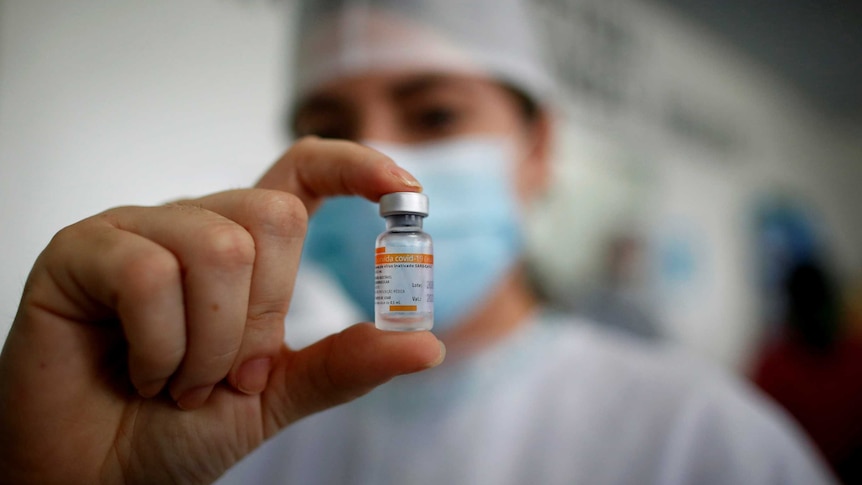 You view a woman holding a tiny vial of the Sinovac vaccine between her thumb and index finger in the centre of the frame.