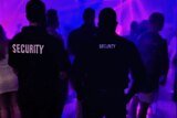Two men with their backs to the camera, with Security written on black shirts, standing in a nightclub.