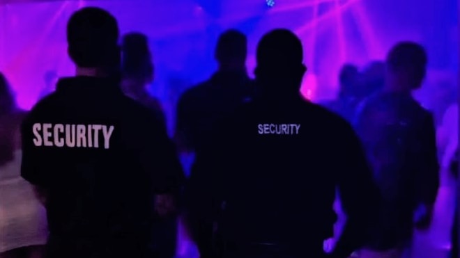 Two men with their backs to the camera, with Security written on black shirts, standing in a nightclub.