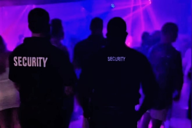 Two men with their backs to the camera, with Security written on black shirts, standing in a nightclub.