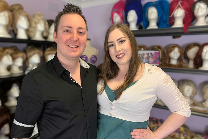 Emily and her partner Rikky stand in front of shelves of wigs.