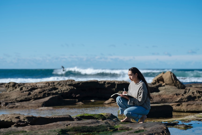 Exteriror scene an artist at the beach drawing with surfer in distance