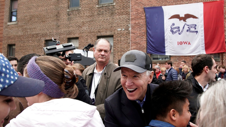 Joe Biden hugs his supporters in a black trucker's cap in front of a brick building with a large Iowan flag draped on it.