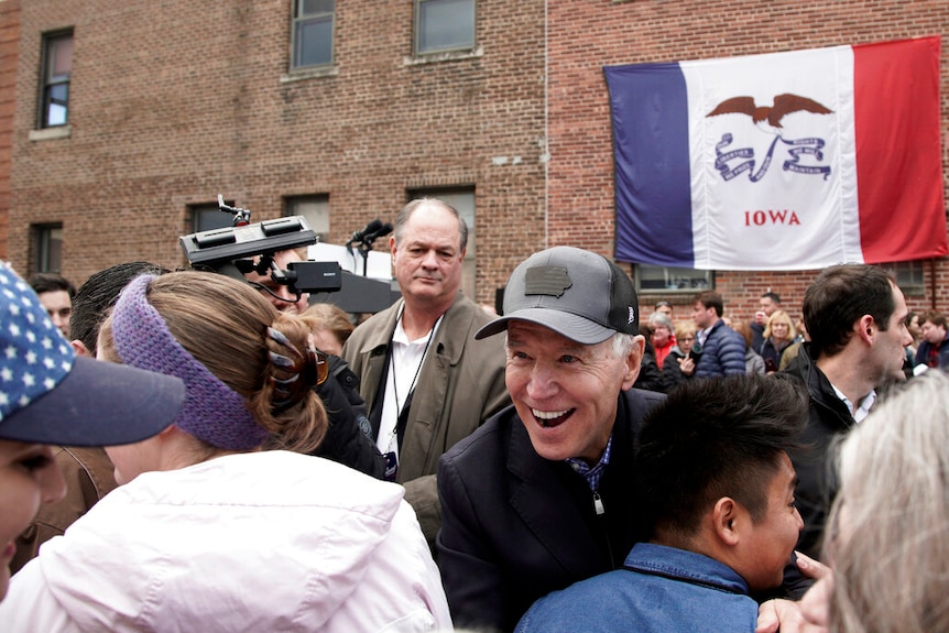 Joe Biden hugs his supporters in a black trucker's cap in front of a brick building with a large Iowan flag draped on it.