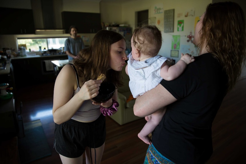 A teenage girl eats a wheatbix while kissing a baby her mum is nursing.