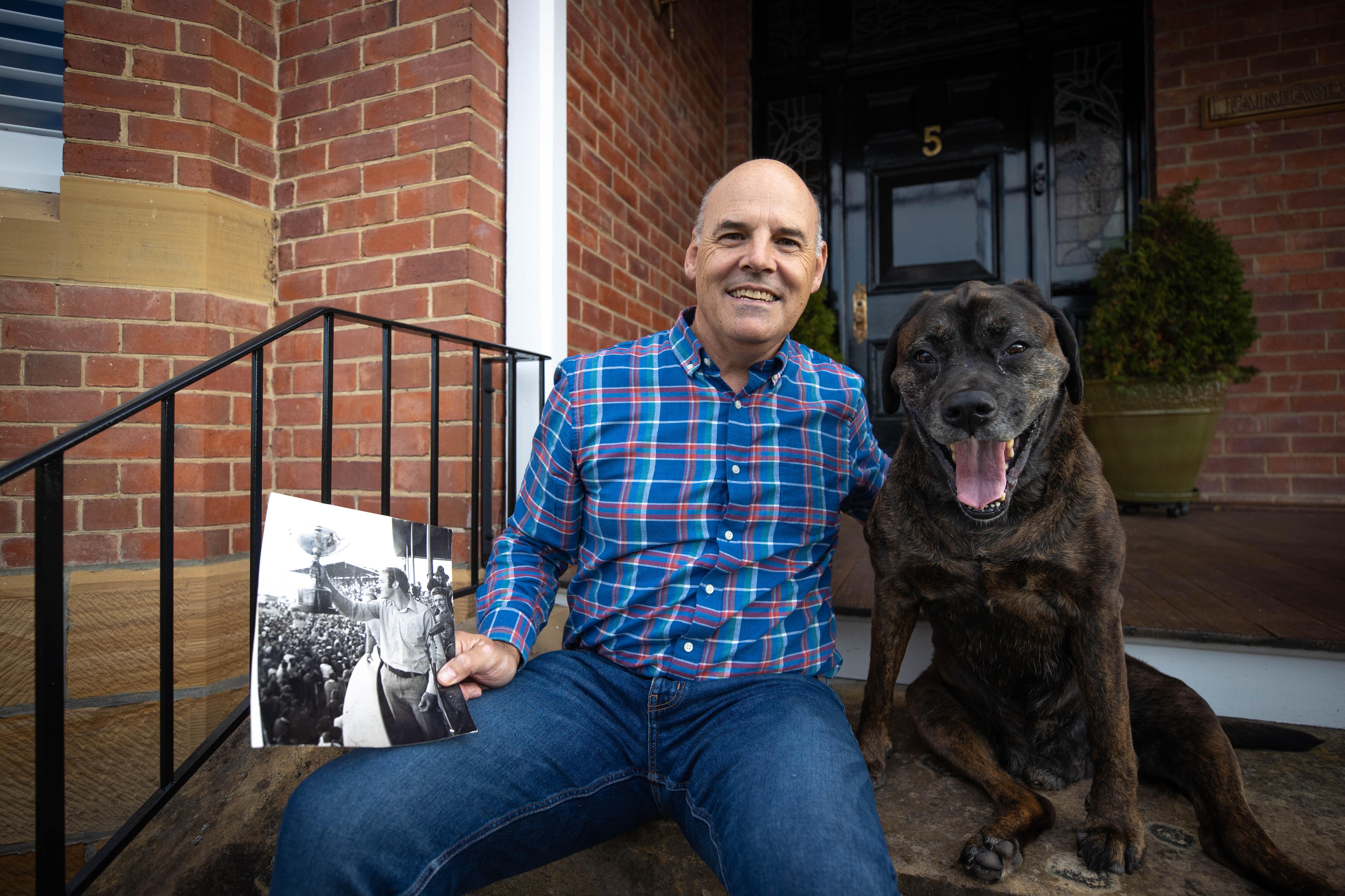 Man in blue checkered shirt sitting in front of house next to his brown dog and holding an old photograph