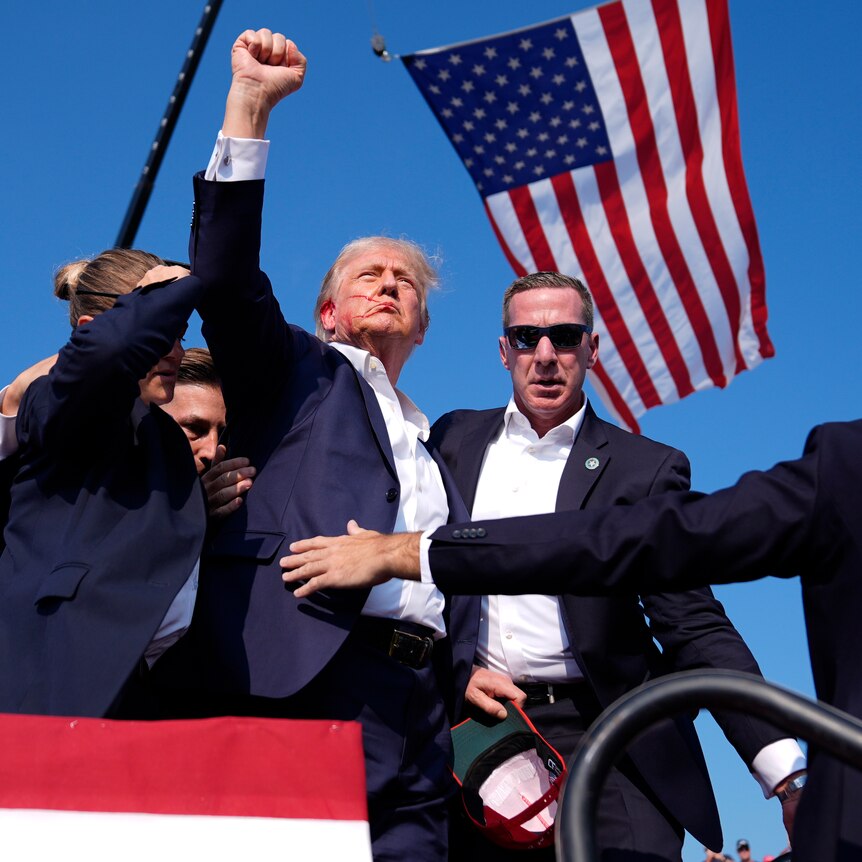 An older man in a suit with blood on his face raises a fist as he's surrounded by Secret Service agents and a US flag waves.