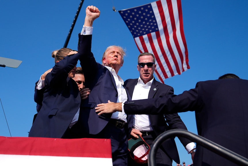 An older man in a suit with blood on his face raises a fist as he's surrounded by Secret Service agents and a US flag waves.