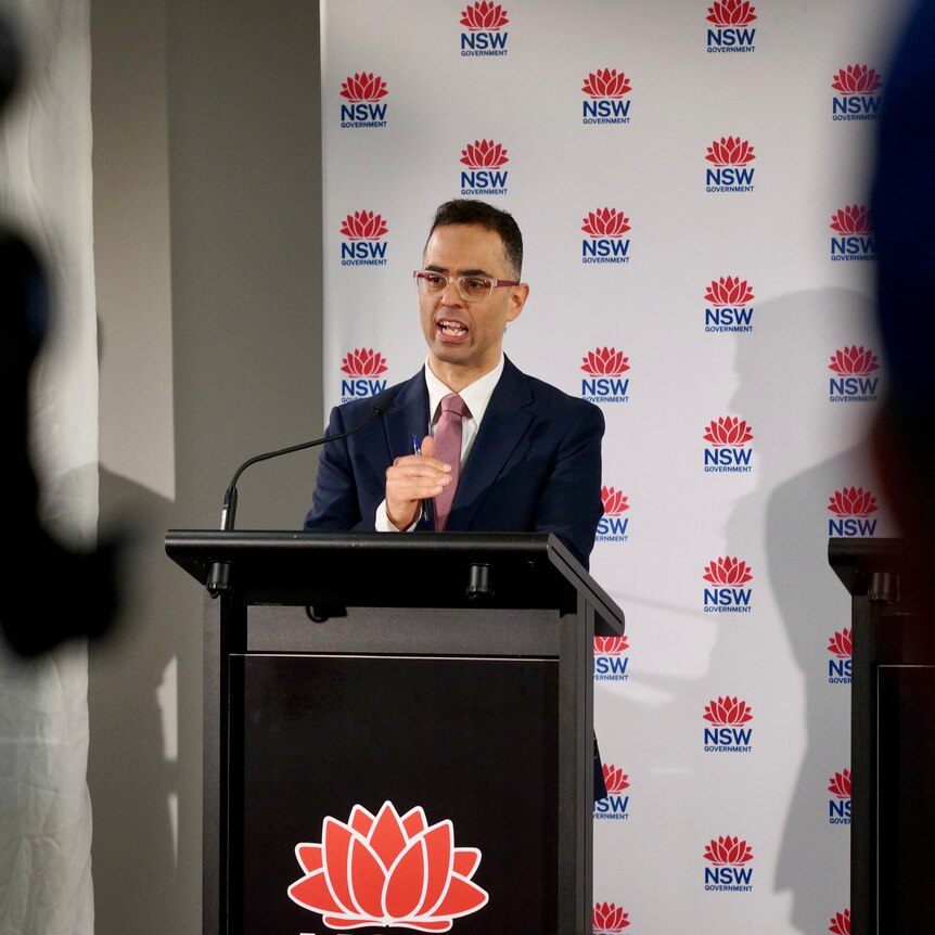 A man in a suit, standing behind a lectern, speaking to reporters at a press conference.