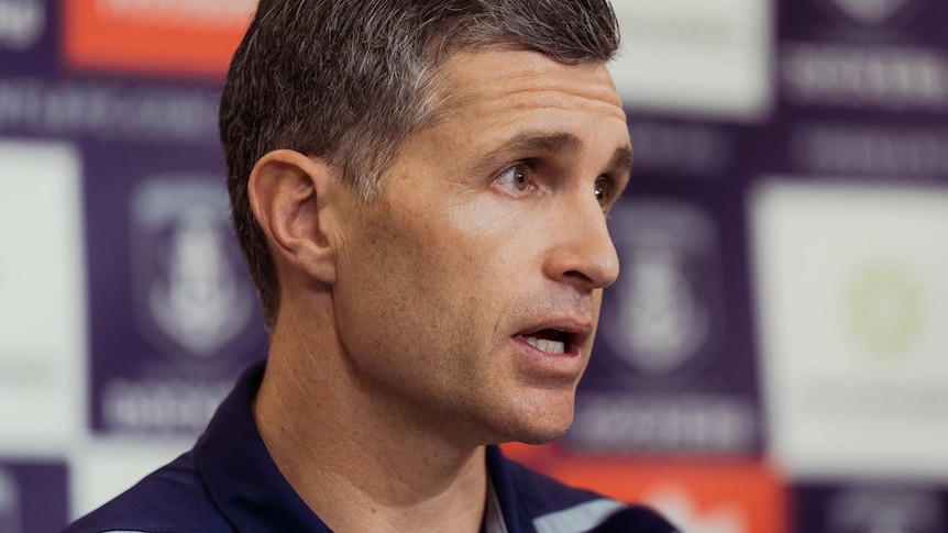 A smiling Justin Longmuir talks during a media conference wearing a purple Fremantle Dockers shirt.