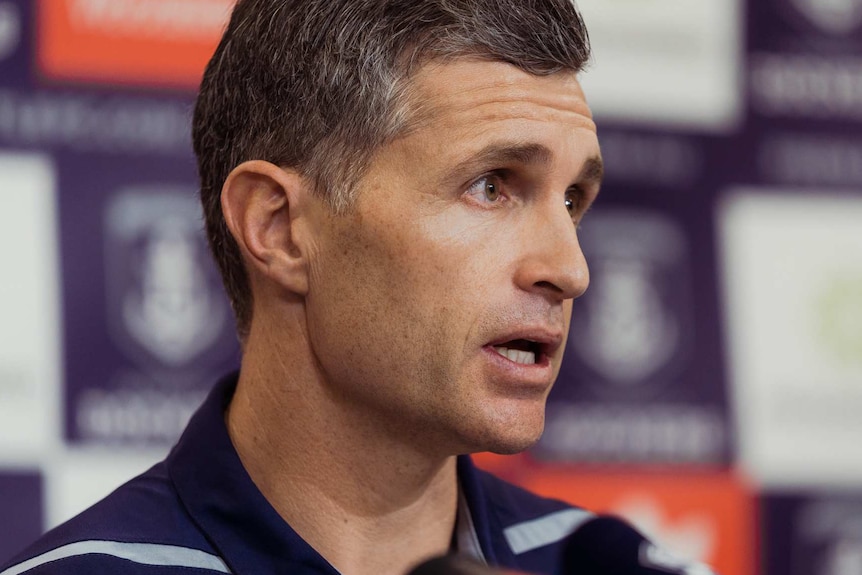 A smiling Justin Longmuir talks during a media conference wearing a purple Fremantle Dockers shirt.