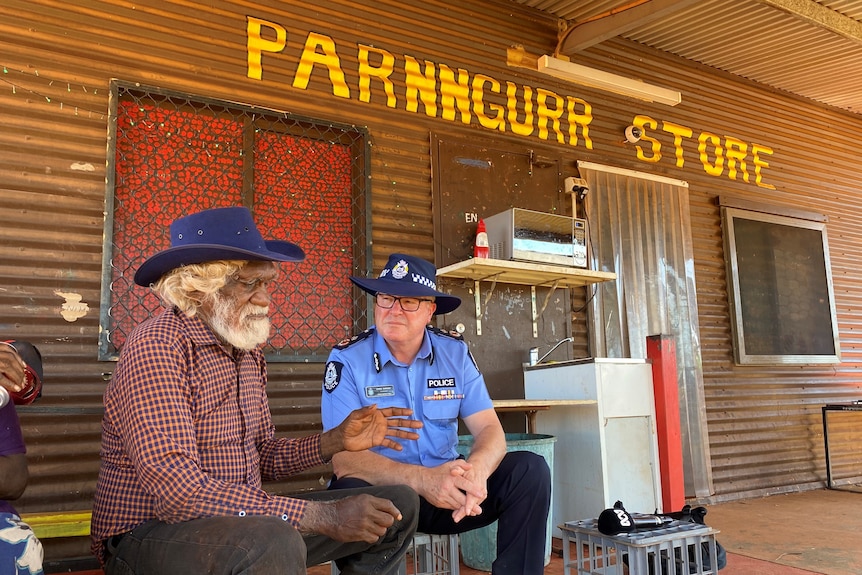 An Indigenous elder wearing hat and checked shirt sits talking with a police officer outside a general store.