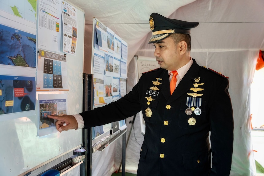 A man in military uniform with orange tie, medals on his navy suit, and a peaked hat, stands next to a whiteboard