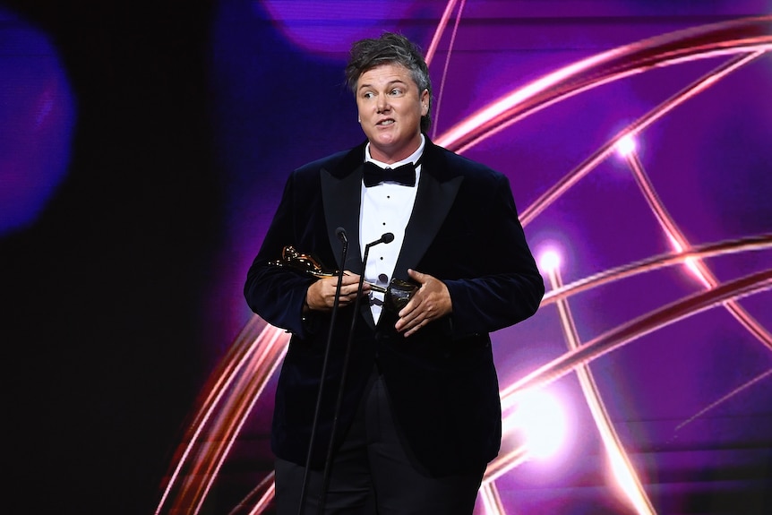 A person with short hair wearing a suit and holding an award with a purple background