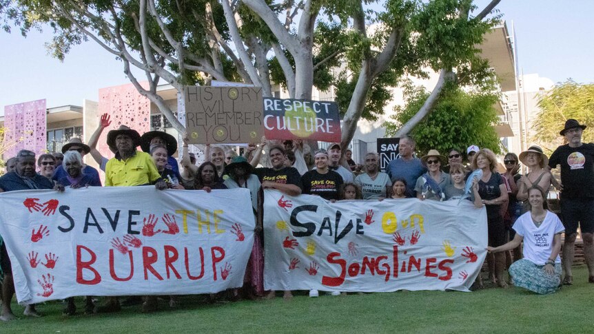 Aboriginal and white people standing on the grass, holding a sign saying Save the Burrup and Save our Songlines