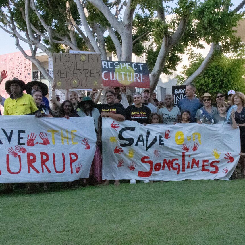 Aboriginal and white people standing on the grass, holding a sign saying Save the Burrup and Save our Songlines