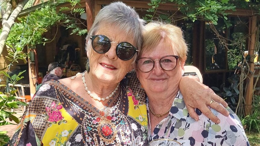 Two senior women stand with hand around each others' shoulders.