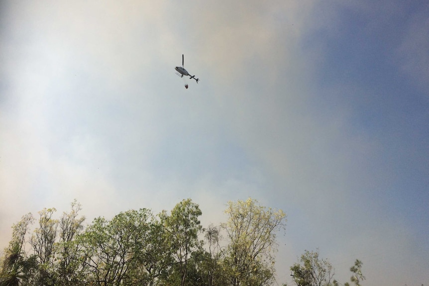 Helicopter with water bucket flies over bushland with heavy smoke rising from trees.