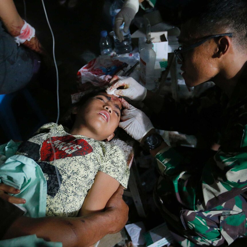 An army doctor examines an injured child outside. The child is lying down, and has a head wound.
