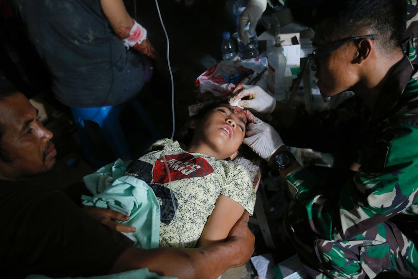 An army doctor examines an injured child outside. The child is lying down, and has a head wound.