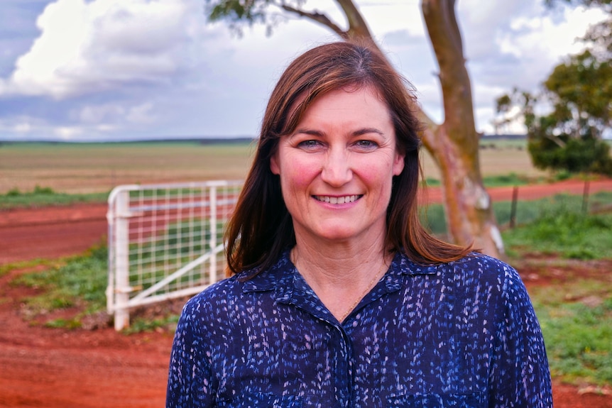 A woman wearing a blue shirt stands in front of a gate with trees in the background.