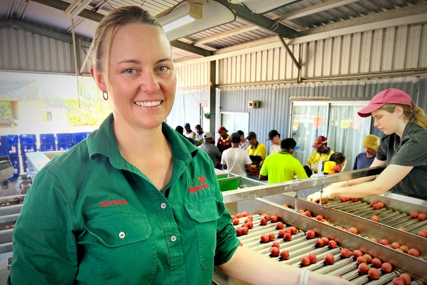 A young woman, in a brightly coloured uniform with the Lush Lychees logo on it, smiles in a busy packing shed