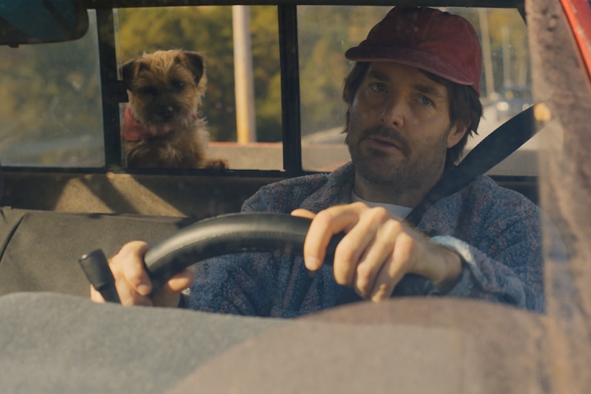 Will Forte as Reggie behind the wheel of a car with a dog in the background. Doug looks exasperated