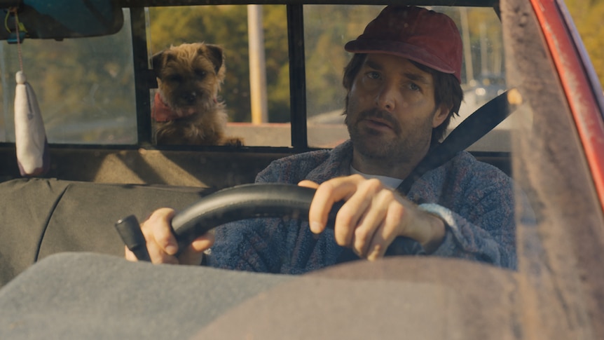 Will Forte as Reggie behind the wheel of a car with a dog in the background. Doug looks exasperated