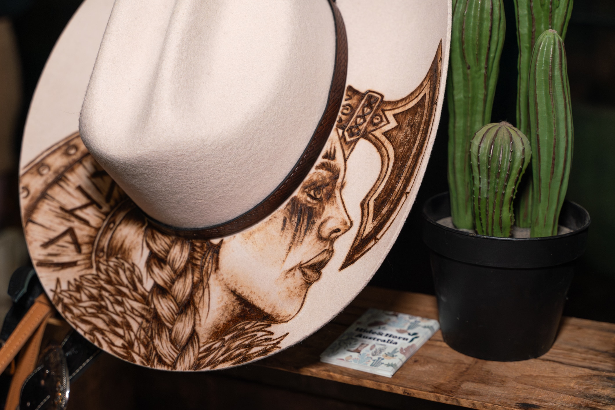 Sombrero de vaquero con cara de mujer y un hacha dibujada en el borde junto a una maceta con cactus